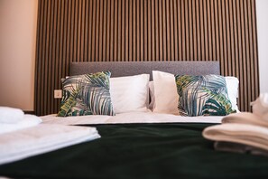 Un lit bien fait, avec une tête de lit grise, des oreillers à motifs blancs et verts, et une couvre-lit vert foncé.