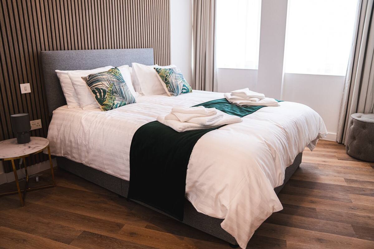 Una cama bien hecha con sábanas blancas y una manta verde, una mesita de noche con lámpara, y piso de madera.