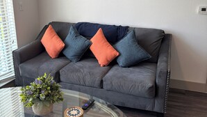 A living room with a grey sofa, blue and orange cushions, a glass coffee table with a plant, and a painting of a bull on the wall.