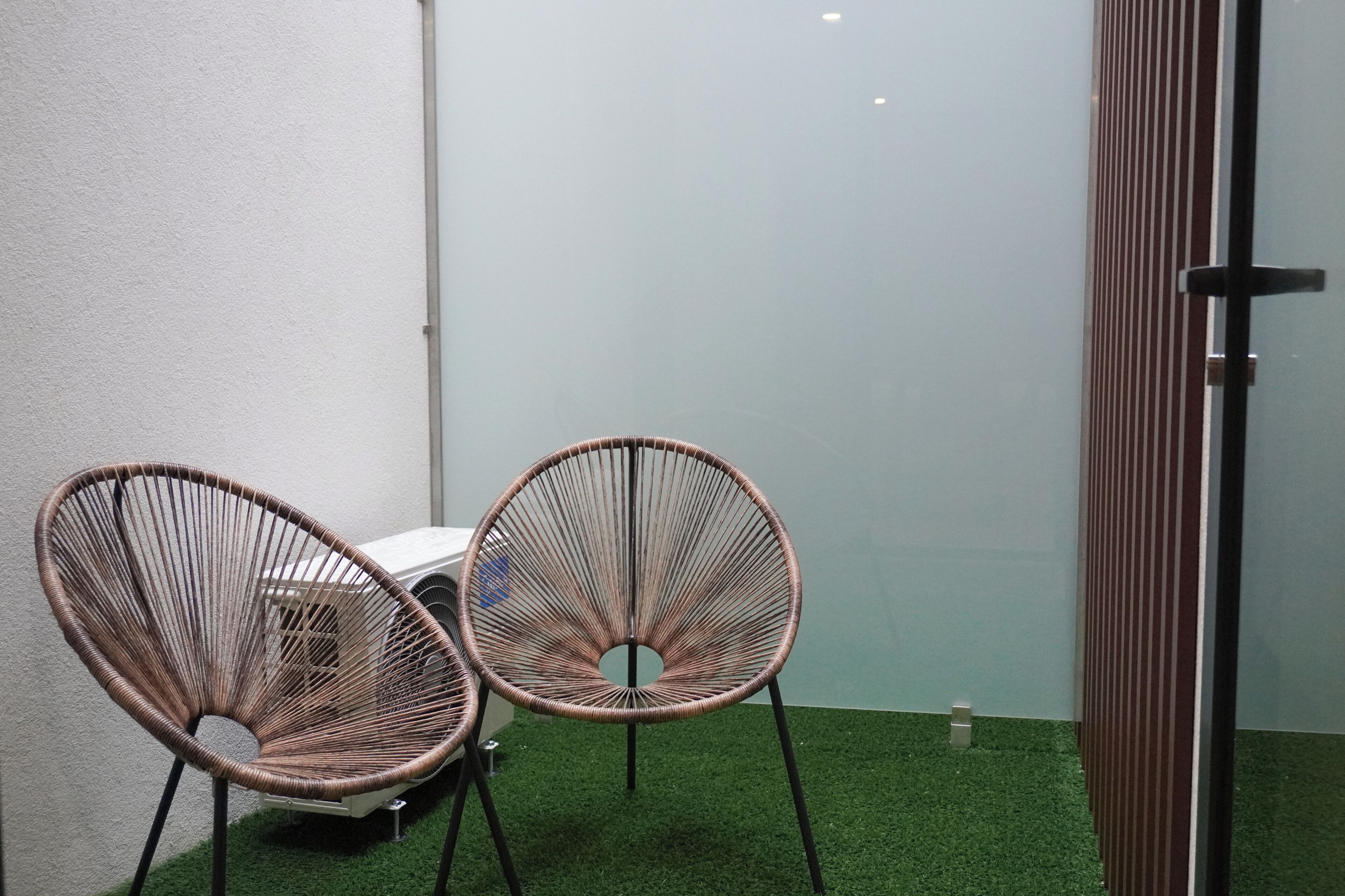 Two wicker chairs with metal legs, a white table, and a glass partition in a room with artificial green flooring.