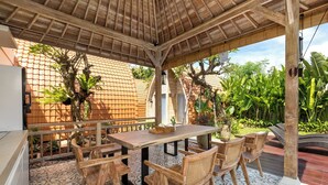 A covered outdoor dining area with a wooden table and chairs, surrounded by patterned tiles and lush greenery. - omaneil (Pecatu)