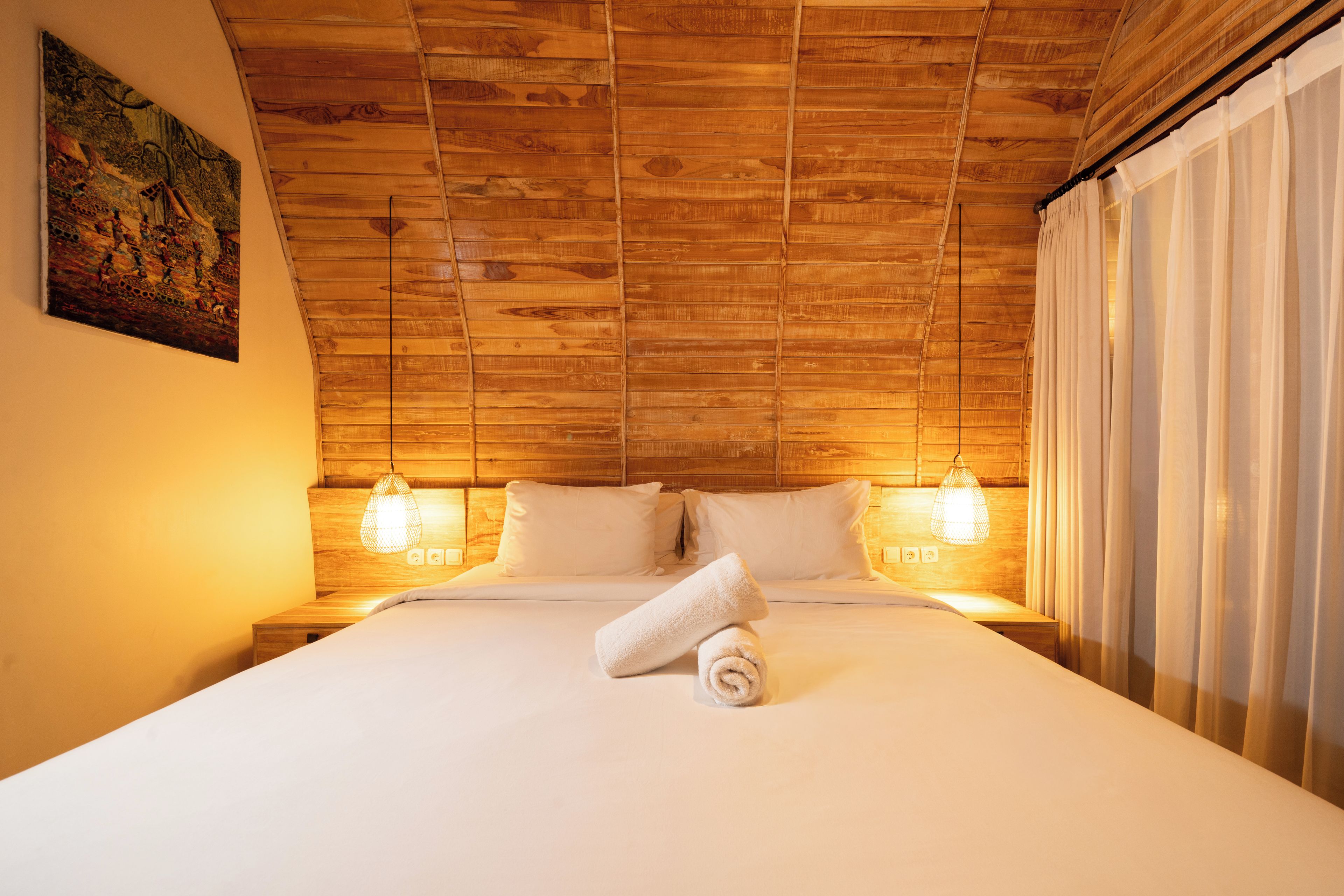 A hotel room with a wooden ceiling, two bedside lamps, a bed with white linens, and a rolled towel on the bed.