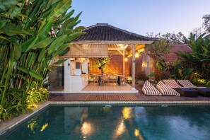 Un área junto a la piscina con zona de estar cubierta, piscina y abundante vegetación.