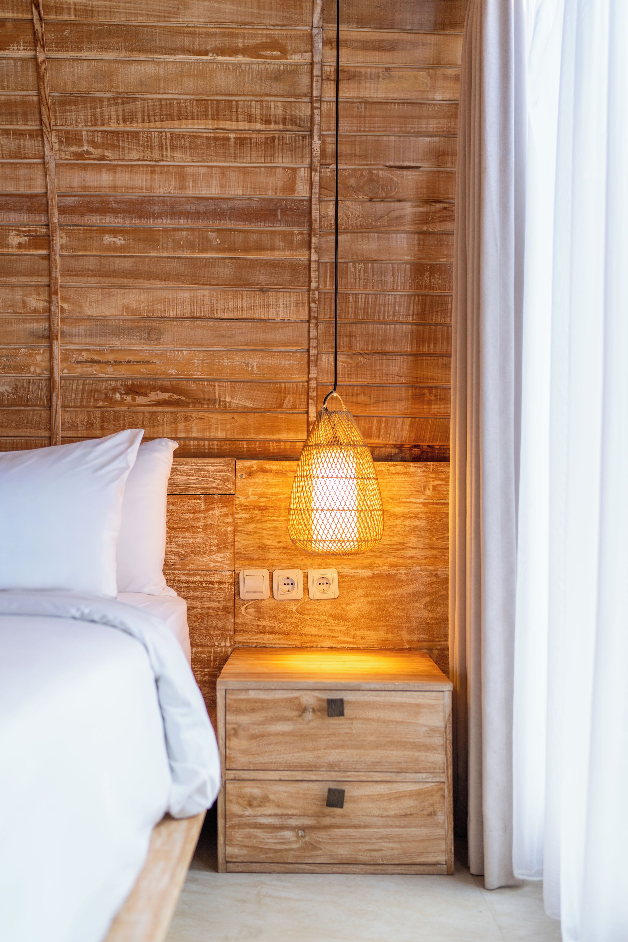 A wooden bed with white bedding, a bedside table with a lamp, and a power outlet.