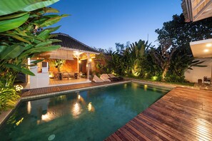 A swimming pool with wooden decking, surrounded by tropical plants and a covered seating area with cushions. - omaneil (Pecatu)
