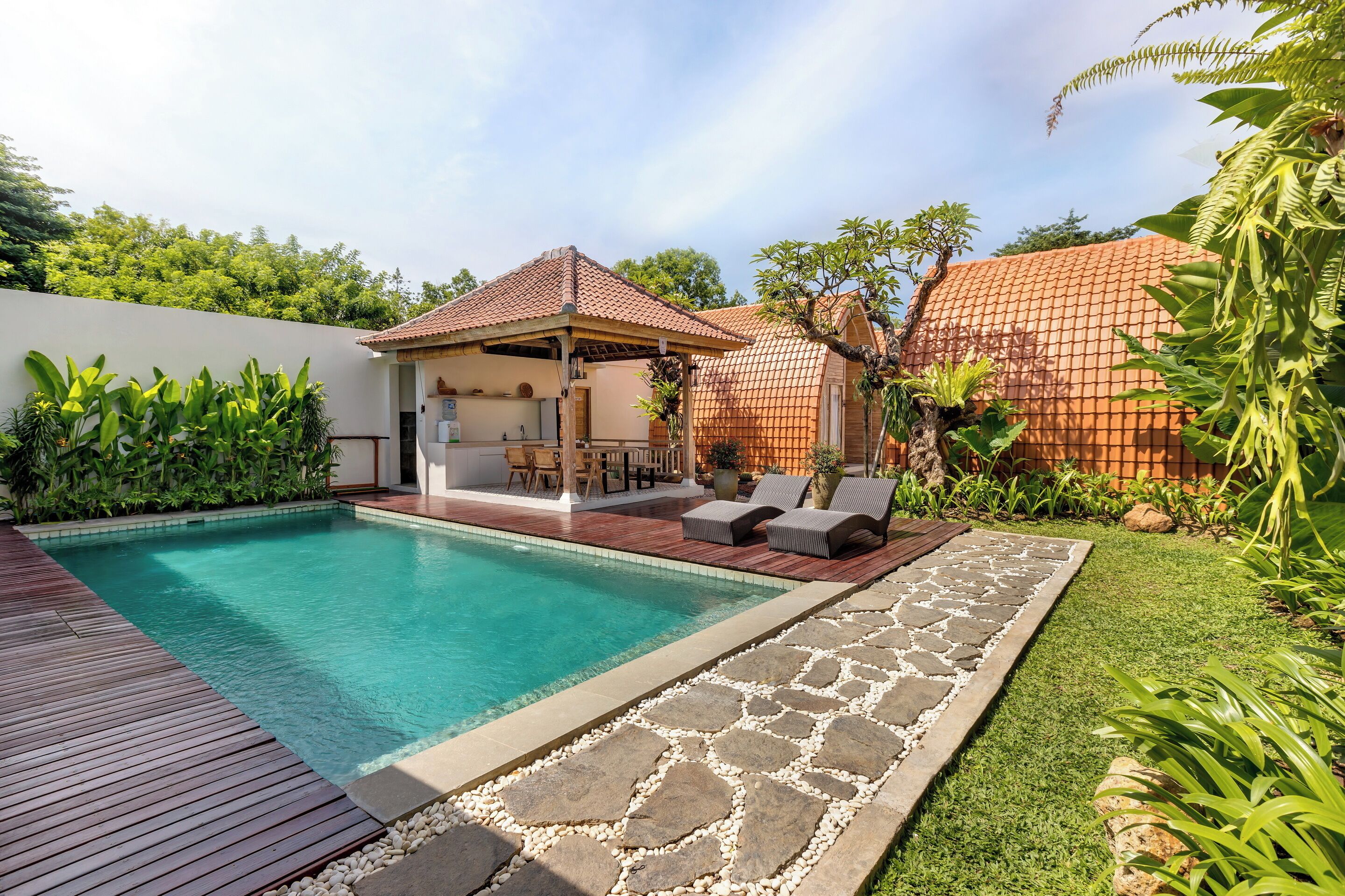 A swimming pool with a wooden deck, stone pathway, and a covered seating area with outdoor furniture.