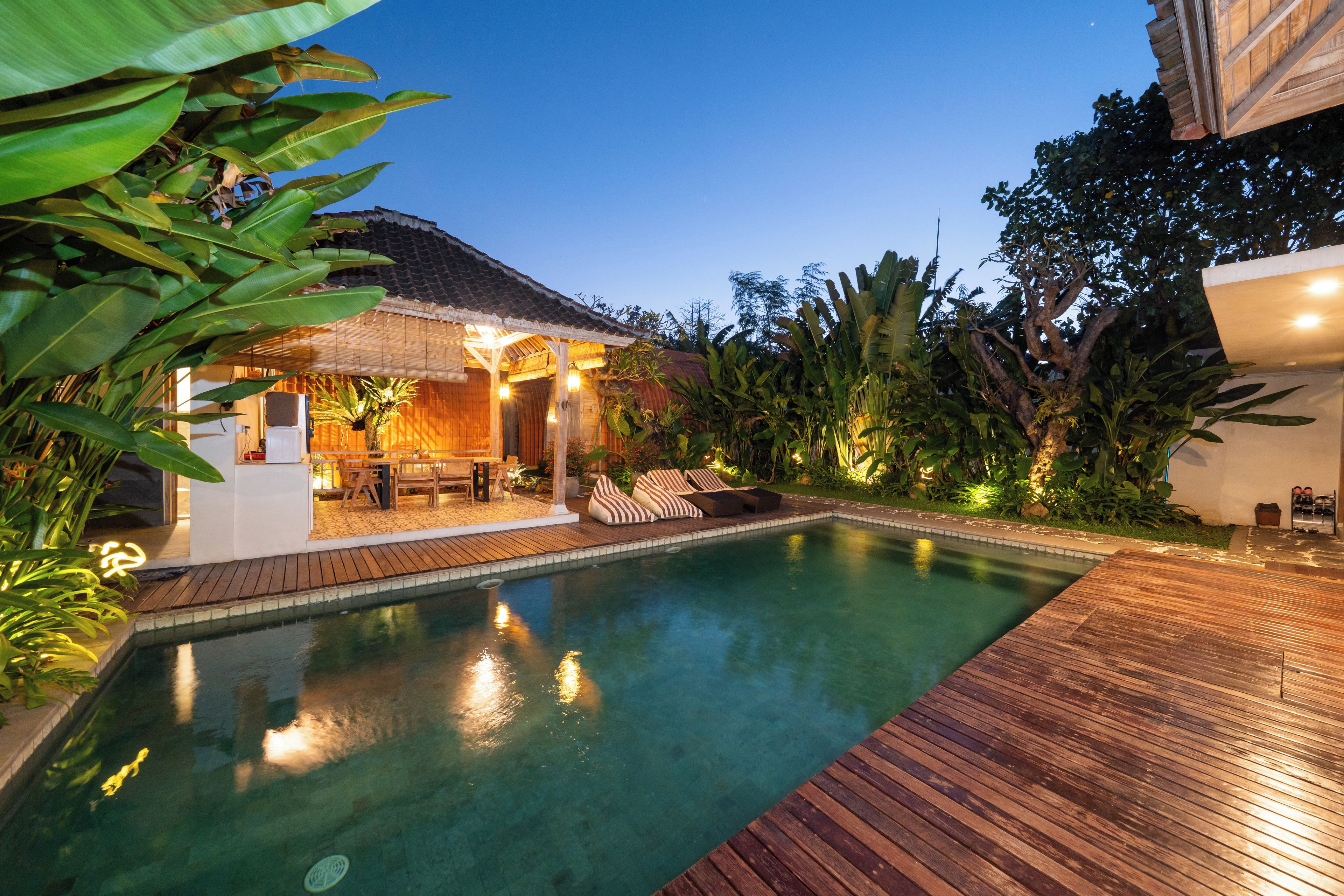 A swimming pool with wooden decking, surrounded by tropical plants and a covered seating area with cushions.