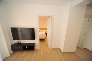 A hotel room with a flat-screen TV mounted on the wall, a wooden floor, and a door leading to a bedroom.