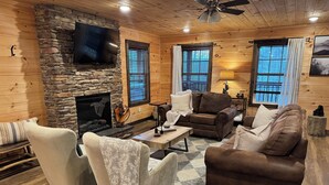 A cozy cabin-style living room with a stone fireplace, wooden walls, and large windows. - Tranquil Haven - Modern Cabin Retreat - Book Your Spring Break Now (Sevierville)