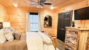 A wooden cabin bedroom with a bed, a dresser, a mirror, a ceiling fan, and a TV. - Tranquil Haven - Modern Cabin Retreat - Book Your Spring Break Now (Sevierville)