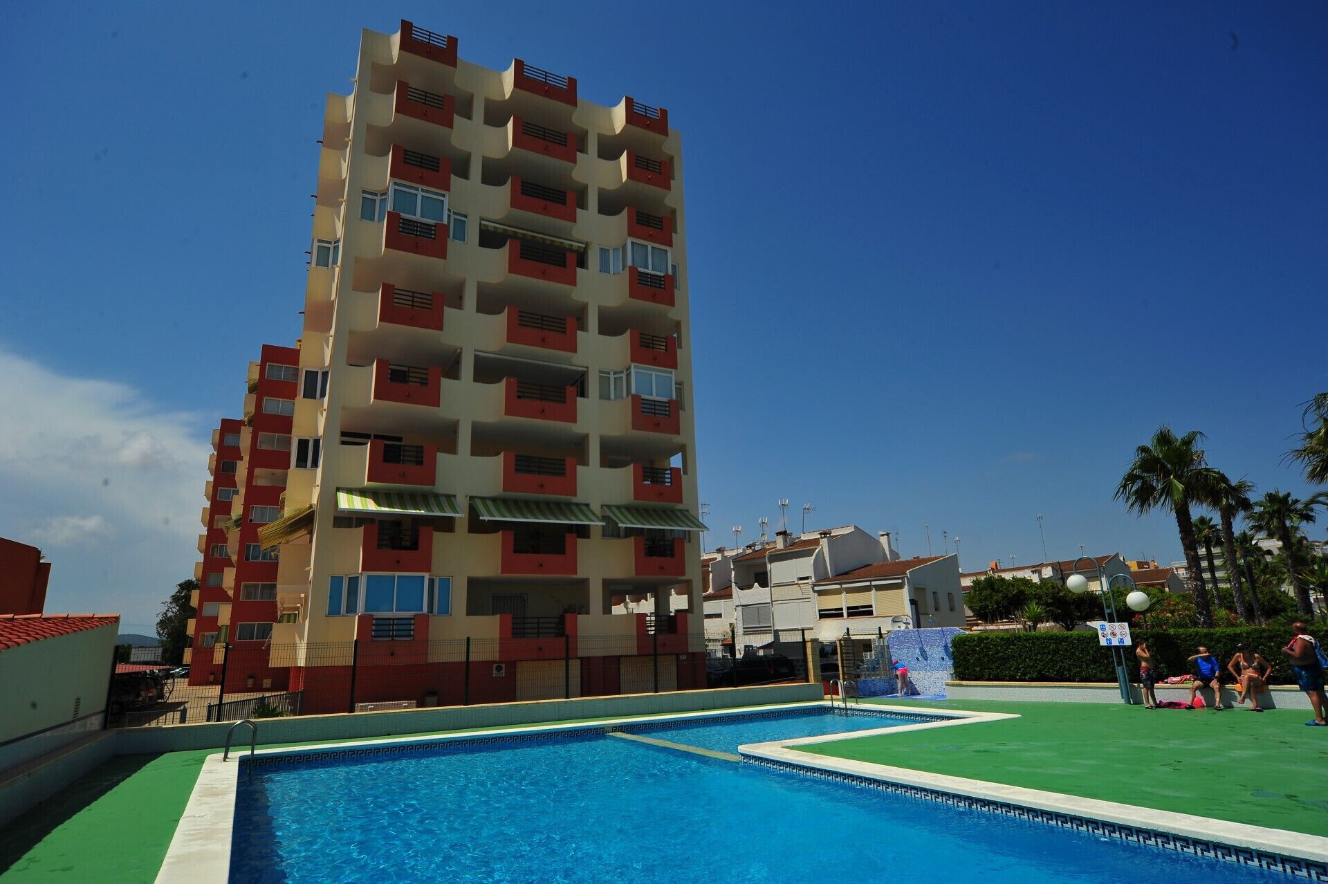 A multi-story hotel with balconies, a swimming pool, and a clear blue sky.