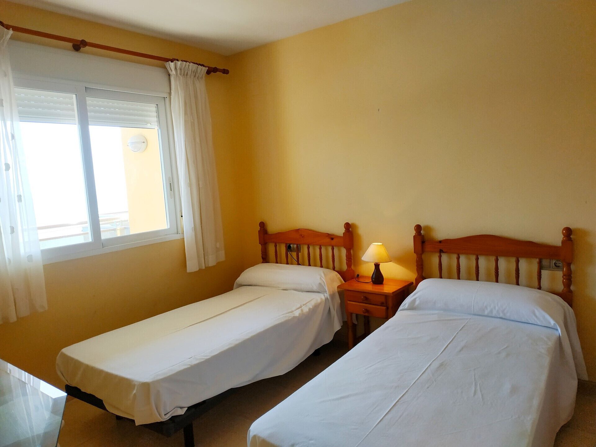 A room with two single beds, a small bedside table with a lamp, a window with curtains, and a wall clock.