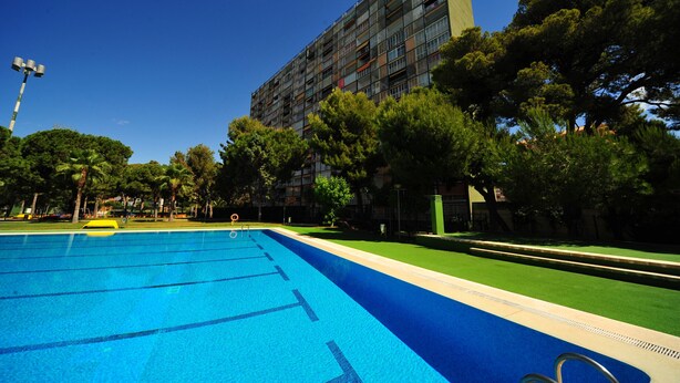 A swimming pool with clear blue water, surrounded by trees and a multi-story building in the background. - PRINCICASIM (Benicàssim)