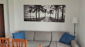 Een woonkamer met een grijze bank, twee blauwe kussens, een houten eettafel en stoelen. Op de muur is een zwart-wit afbeelding van een strand te zien.