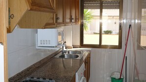 A kitchen with wooden cabinets, a stainless steel sink, a stove, a washing machine, and a window with a view of the outside. - UPPER SHORE (Alcoceber / Alcossebre)