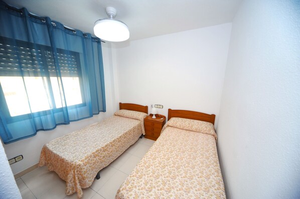 A small, clean room with two single beds, a nightstand, a lamp, and a window with blue curtains.