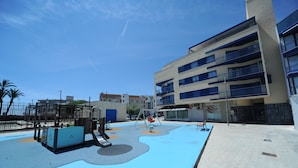 Un parque infantil con suelo azul y columpios, junto a un edificio de varios pisos con balcones.