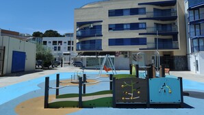 Un parque infantil con diversas estructuras de juego, un edificio con múltiples balcones y un cielo azul despejado.