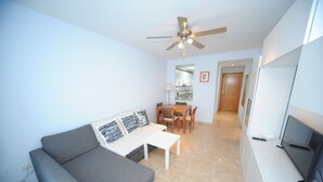 A compact living space with a grey sofa, a small dining table, a ceiling fan, and a TV on a white cabinet. - BAIX MAESTRAT (Peñiscola)