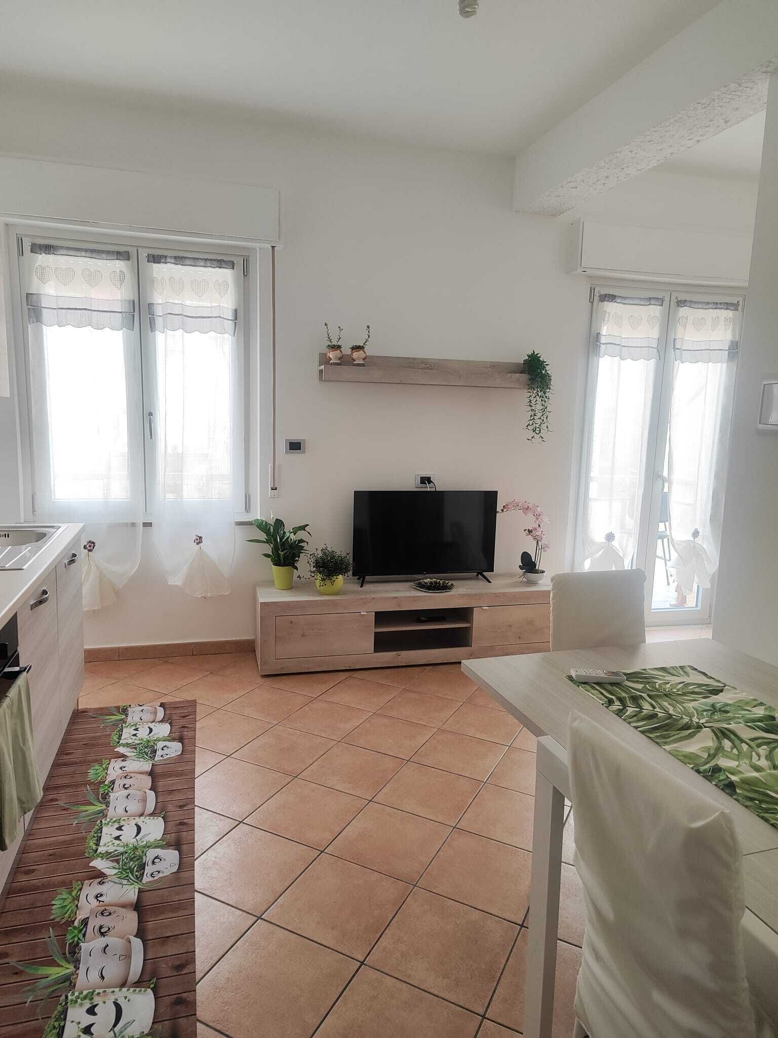 Un soggiorno moderno con una TV a schermo piatto su un mobile in legno, una zona pranzo con tavolo e sedie, e un angolo cottura con lavello e pensili.