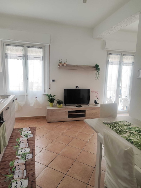 A modern living room with a flat-screen TV on a wooden stand, a dining area with a table and chairs, and a kitchen area with a sink and cabinets.