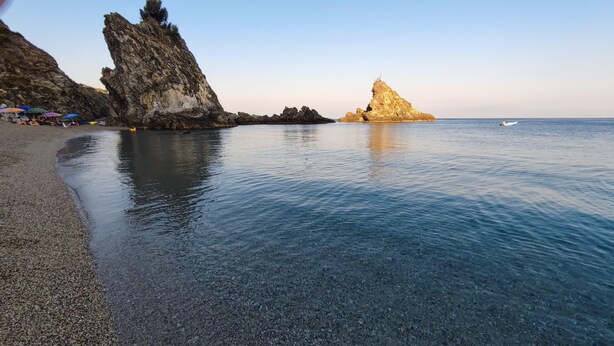 A pebble beach with clear water, rocky formations, and a distant boat. - Palmi Bay Home (Palmi)