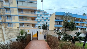 A pathway leading to a hotel entrance with a blue and beige facade, surrounded by greenery and a clear sky.