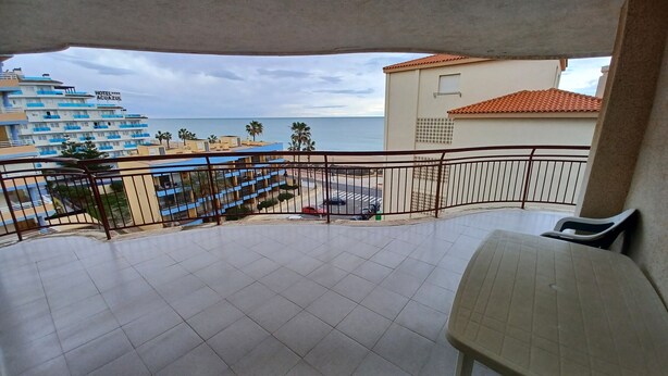 A balcony with a view of the sea, a building with a blue and white facade, and a red car parked on the street.