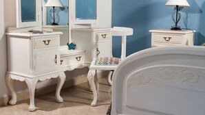 A room with a white bed, a white dresser with drawers, a mirror, and a nightstand with a lamp.