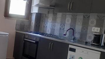 La cuisine est équipée d’une cuisinière à gaz, d’un four et d’un lave-vaisselle. On y trouve une fenêtre avec des stores, un évier avec un robinet violet et une machine à café posée sur le plan de travail.