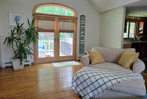 Una sala de estar con piso de madera, un sofá color beige con cojines amarillos, una planta grande en maceta y una puerta que da a un espacio exterior.