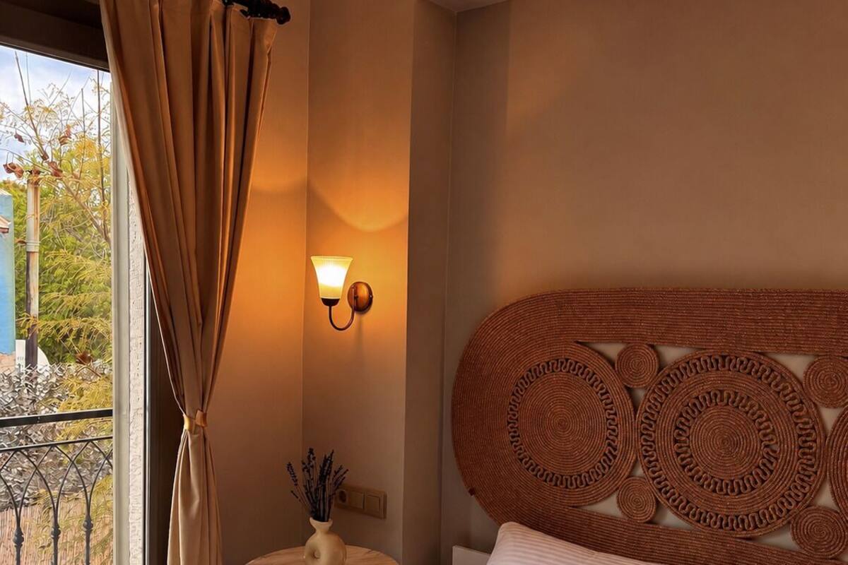 A hotel room with a bed, a small table with a vase, a wall-mounted lamp, and a view of the outside.