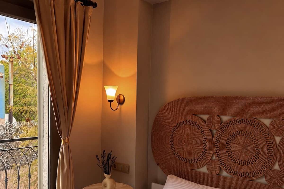A hotel room with a bed, a small table with a vase, a wall-mounted lamp, and a view of the outside.