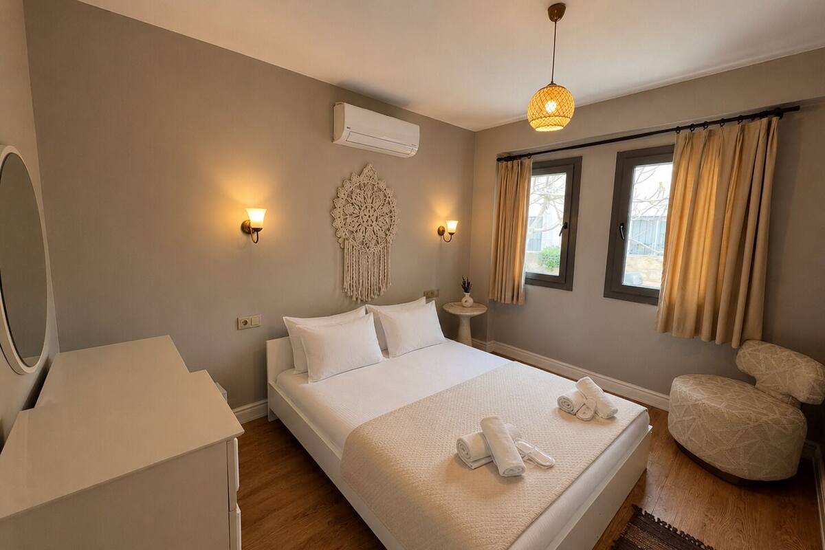 A bedroom with a bed, bedside lamps, a wall-mounted air conditioner, a decorative wall hanging, a small table with a plant, and a window with curtains.