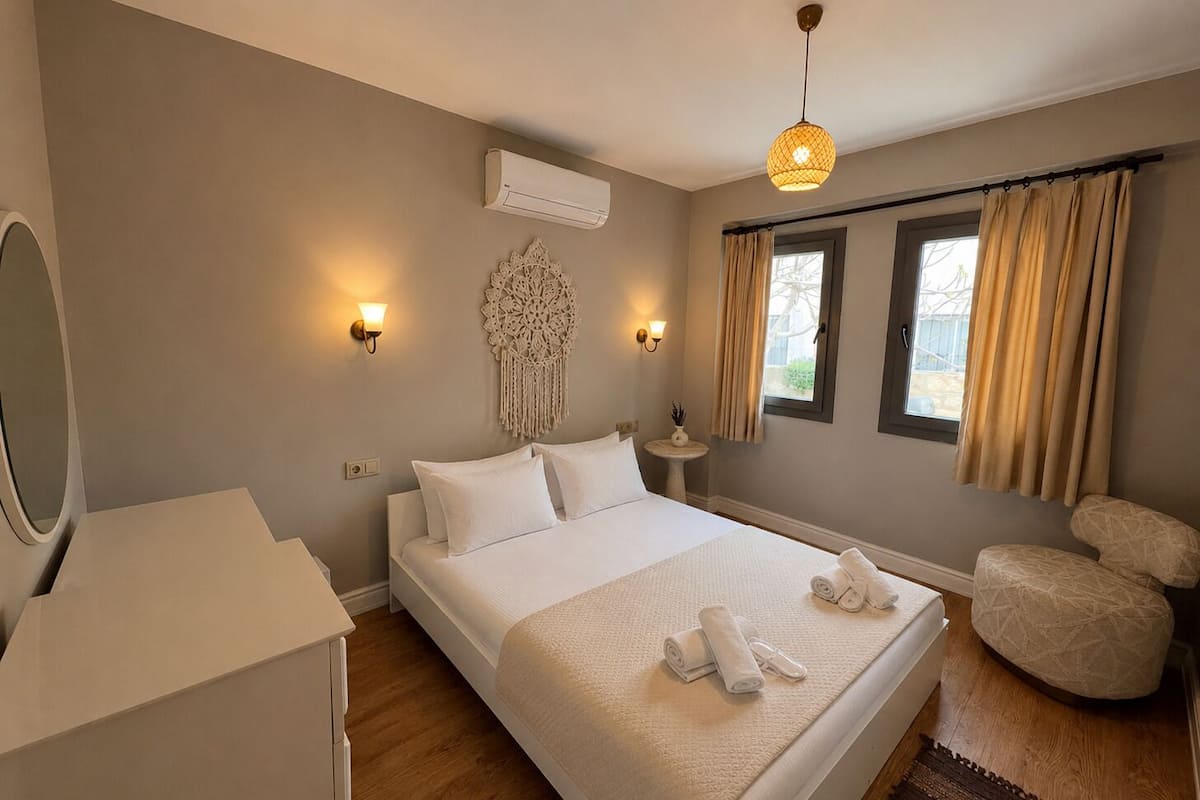 A bedroom with a bed, bedside lamps, a wall-mounted air conditioner, a decorative wall hanging, a small table with a plant, and a window with curtains.