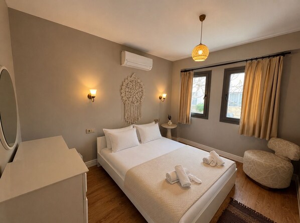 A bedroom with a bed, bedside lamps, a wall-mounted air conditioner, a decorative wall hanging, a small table with a plant, and a window with curtains. - No9 Alacati (Cesme)