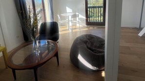Een moderne woonkamer met een glazen salontafel, een zwarte stoel en een relaxfauteuil. Er is een balkon met uitzicht op bomen en een deur naar buiten.