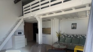 Een moderne loft-achtige leefruimte met een hoge plafond, een trap naar een hoger niveau, een eethoek met een ronde tafel en stoelen, een bank en een klein keukentje.