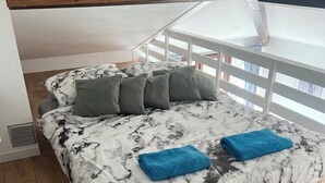 Een bed met een sprei met marmerpatroon, twee blauwe handdoeken en meerdere kussens in een kamer met houten vloeren en een witte leuning.