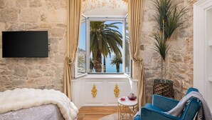 Une pièce avec un mur en pierre, une grande fenêtre avec des rideaux, un téléviseur à écran plat, une chaise bleue, une petite table ronde avec une théière, et un plaid en fausse fourrure blanche sur le lit.