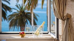 Une fenêtre donnant sur des palmiers et l’océan, une bouteille de champagne, deux verres et un bol de fraises posés sur le rebord de la fenêtre.