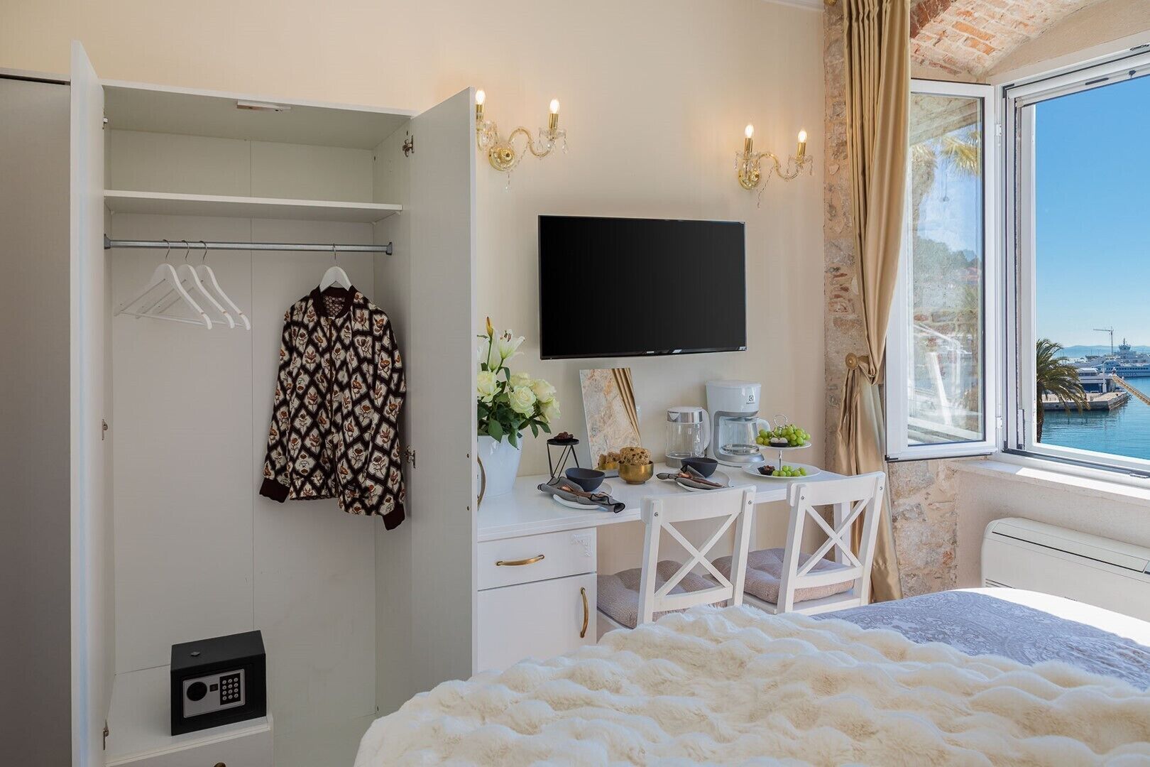 A bedroom with a bed, a desk with a computer, a TV, a wardrobe, and a window with a view of a marina.