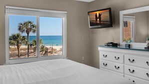 Een slaapkamer met een groot raam met uitzicht op een strand, een bed, een dressoir en een televisie aan de muur.