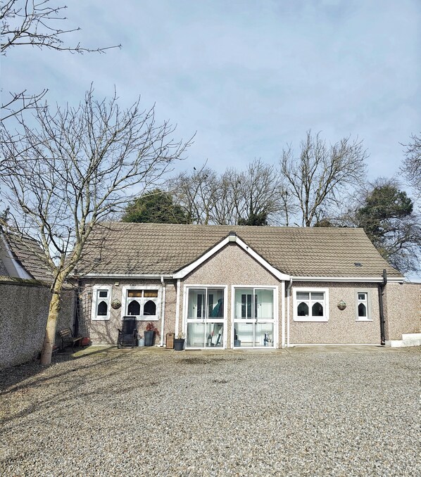 A two-story building with a tiled roof, a gravel driveway, and a stone wall. - The Curragh Studio (Cut Bush)