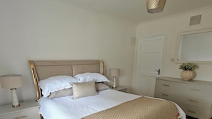 A bedroom with a large bed, bedside tables, a dresser, a mirror, and a hanging light fixture. - The Curragh Studio (Cut Bush)