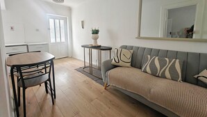 A modern living room with a grey sofa, a round wooden dining table with chairs, a small side table with a plant, and a large mirror on the wall. - The Curragh Studio (Cut Bush)