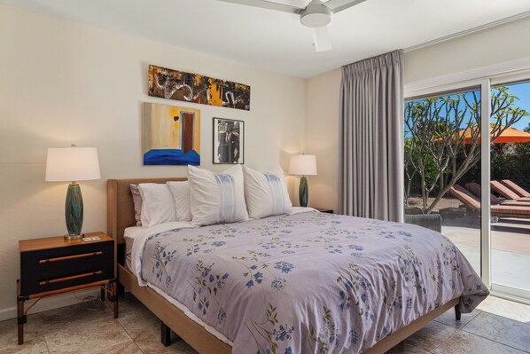 A hotel room with a bed, two bedside tables, a ceiling fan, a large window with curtains, and artwork on the wall.
