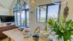A dining area with a wooden table set for a meal, a TV in the background, and large windows offering a view of the outdoors. - 2 Trem Y Mor -  a dog that sleeps 6 guests  in 3 bedrooms (Moelfre)