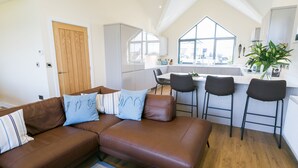 A modern living room with a brown leather sofa, a dining area with a white table and black chairs, and a kitchen with white cabinets and a large window. - 2 Trem Y Mor -  a dog that sleeps 6 guests  in 3 bedrooms (Moelfre)