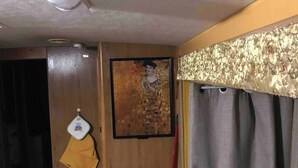 A room with a wooden door, a framed artwork on the wall, a red broom leaning against the wall, a bed with a patterned bedspread, and a small wooden stool. - Gustav Klimt The Kiss -Camper/RV in Saint Matthews, South Carolina (Saint Matthews)